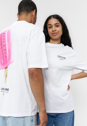 Deux personnes portent des T-shirts blancs Coolture ; l'une montre un grand motif rose de glace à l'eau dans le dos, l'autre a un petit texte devant.