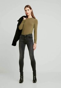 Top de manga longa em oliveira combinado com jeans skinny pretos de cintura alta. O modelo usa botas pretas até o tornozelo e segura uma jaqueta preta sobre um ombro.