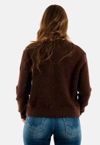 Maglione lavorato a maglia marrone con superficie testurizzata, caratterizzato da macchie colorate e orlo a coste, abbinato a jeans in denim blu con un design unico delle tasche.