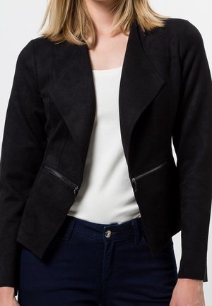 Frau trägt eine eng anliegende schwarze Jacke mit diagonalen Reißverschlusstaschen über einem weißen Oberteil und dunkelblauen Jeans, gezeigt von den Schultern bis zu den Hüften.