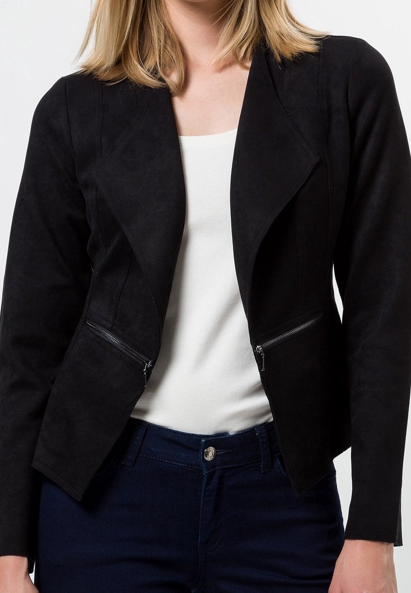 Femme portant une veste noire ajustée avec des poches zippées en diagonale par-dessus un haut blanc et un jean bleu foncé, montrée des épaules aux hanches.