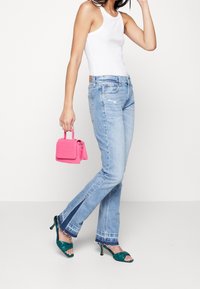 Jeans de corte recto en un tono azul claro con efecto desgastado y aberturas laterales, combinados con una camiseta sin mangas blanca y un bolso rosa. Sandalias de tacón verdes.