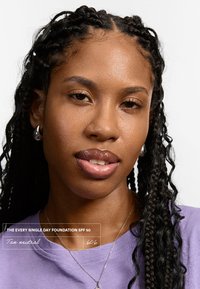 Beżowy neutralny podkład w butelce, nałożony na skórę. Osoba ma zaplecioną fryzurę, nosi fioletową koszulę i srebrne kolczyki w kształcie kółek. Gładka tekstura skóry.