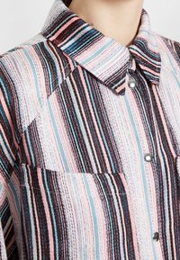 Chemise rayée colorée avec un col à boutons, boutons en argent et une poche poitrine. Le tissu présente un motif texturé multicolore.