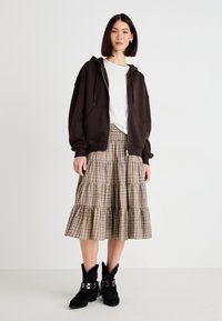 Woman wearing a dark brown zip-up hoodie, white t-shirt, beige plaid tiered skirt, and black ankle boots standing against a plain white background.