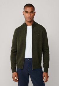 Olijfgroene ritscardigan van zachte stof, met een hoge kraag en geribde manchetten, gedragen over een wit T-shirt en marineblauwe broek.