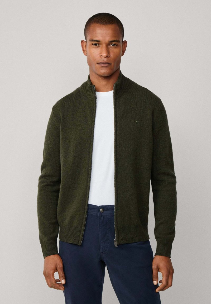 Olijfgroene ritscardigan van zachte stof, met een hoge kraag en geribde manchetten, gedragen over een wit T-shirt en marineblauwe broek.