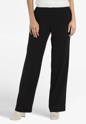 Vrouw in zwarte wijde broek en witte enkellaarsjes, handen naast de heupen, staand op een witte achtergrond.