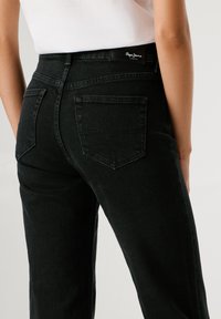 Fekete farmernadrág magas derékkal, két hátsó zsebbel, amelynek varrott mintázata van. Az anyagnak enyhe textúrája van és tiszta befejezése.