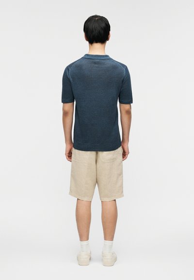 Mørkeblå kortermet polotrøye laget av mykt stoff, kombinert med lyse beige linsjorts. Modellen står med ryggen til for å vise frem klærne.