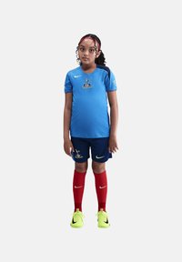 Maillot de football Nike bleu avec un logo, accents bleu marine, associé à un short foncé et des chaussettes rouges. Chaussures vert vif avec des détails noirs.