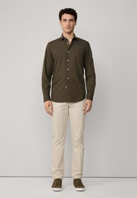 Camisa de botones de color verde oliva oscuro, de manga larga, con una tela suave, que presenta botones blancos y un sutil acento de logo. Combinada con pantalones beige y zapatos verdes.