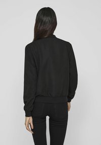 Zwarte bomberjack met lange mouwen, geribbelde manchetten en zoom, en een gladde stoftextuur. Draagt zwarte nauwsluitende broek, gezien vanaf de achterkant.