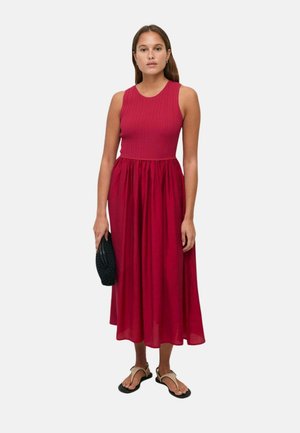 Mujer con vestido midi rojo sin mangas con parte superior acanalada y falda fruncida, llevando un bolso negro y sandalias beige.