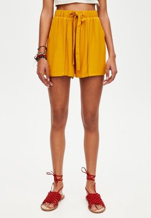 Femme portant un short taille haute moutarde avec une ceinture nouée, un haut court blanc, des sandales rouges tressées à lacets et des bracelets au poignet gauche.
