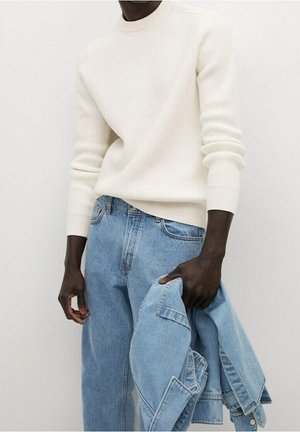 Osoba ubrana w biały prążkowany sweter i jasnoniebieskie dżinsy, trzymająca jasnoniebieską kurtkę dżinsową w lewej ręce na jednolitym tle.