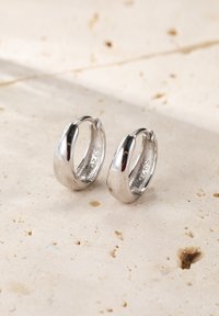 Srebrne kolczyki w kształcie obręczy, gładka powierzchnia, zaokrąglony kształt, mały rozmiar, z widoczną próbą wewnątrz. Na tle teksturowanego kamienia.