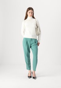 Maglione a collo alto bianco e testurizzato abbinato a pantaloni a sigaretta color teal e décolleté nere a punta, mentre tiene una borsa chiara. Outfit semplice e moderno.