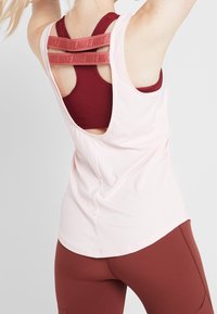 Top sportivo atletico rosa chiaro con schiena a scollatura rotonda, sovrapposto a un reggiseno sportivo color bordeaux con spalline incrociate, realizzato in tessuto traspirante.
