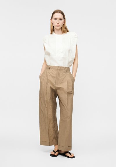 Γυναίκα με ίσια ανοιχτό καφέ μαλλιά, που φοράει λευκό αμάνικο τοπ, μπεζ παντελόνι ψηλόμεσο με φαρδιές παντελόνες και μαύρα σανδάλια, όρθια με τα χέρια στις τσέπες.