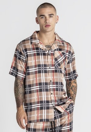 Mladý muž s krátkymi vlasmi, oblečený v béžovej, čiernej, bielej a červenej kockovanej košeli s krátkym rukávom a zodpovedajúcich nohaviciach, ktorý ukazuje tetovania na hrudi a rukách.