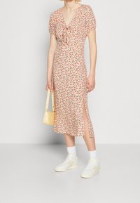 Nainen, jolla on päällään vaaleanpunainen kukallinen midi-mekko puffihihnoilla ja sivuaukolla, valkoiset lenkkarit, valkoiset sukat ja pieni vaaleankeltainen käsilaukku.