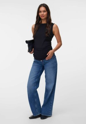 Kvinde iført blå vidbenede jeans og sort ærmeløs top, der holder en sort clutch mod en ensfarvet hvid baggrund.
