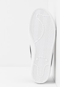 Černé tenisky s bílou gumovou podrážkou s vzorem rybí kosti a bočním švem. Text na podrážce: "VYROBENO VE ŠPANĚLSKU."