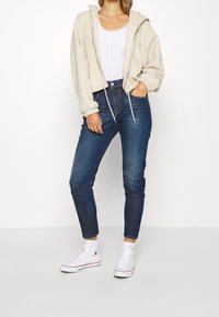 Beige verkürzter Hoodie mit Kordelzug, kombiniert mit dunkelblauen Slim-Fit-Jeans und weißen High-Top-Sneakern. Darunter ein strukturiertes Strick-Top.