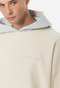 Beige trui met een grijze capuchon, voorzien van een textiele stof en geborduurd "PEGAJOR" logo op de borst. Het ontwerp heeft een ontspannen pasvorm.