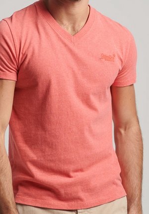 Mand iført en koralrosa t-shirt med korte ærmer og V-hals samt beige bukser mod en ensfarvet lyse baggrund.