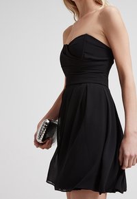 Zwarte strapless jurk met een aansluitend lijfje en een vloeiende rok, gemaakt van lichtgewicht stof. Geaccentueerd met een zilveren clutch in de hand.
