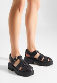 Black platform sandals with an open-toe design, featuring crisscross straps, a buckle accent, and a textured rubber sole.