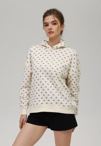 Sudadera con capucha en color crema con un pequeño patrón de lazo morado que se repite. Tela suave, ajuste relajado, y puños y dobladillo acanalados; combinada con pantalones cortos negros.