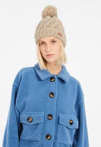 Bonnet tricoté beige avec un pompon sur le dessus, présentant un motif texturé. Veste en polaire bleue avec un col et des boutons noirs, et deux poches sur la poitrine.