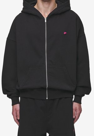 Person iført en sort hættetrøje med lynlås og en lille pink "P" logo på venstre bryst samt matchende sorte bukser.