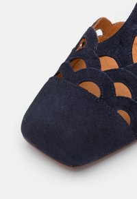Gros plan d'une chaussure plate en daim bleu marine avec un décor festonné découpé et un bout carré sur fond blanc.