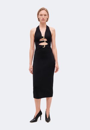 Le mannequin se tient devant un fond uni, vêtu d’une robe midi noire avec des boucles dorées et des escarpins noirs à bout pointu.