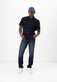 Camicia a maniche corte blu navy con bottoni, jeans blu scuro, sneakers bianche, cappellino azzurro; modello in piedi con le mani nelle tasche, sfondo neutro.