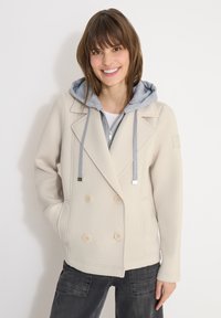 Jeune femme souriante portant une veste beige à double boutonnage avec un sweat à capuche gris en dessous et un jean foncé.