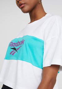 Bílý bavlněný crop top s tyrkysovým vodorovným pruhem. Obsahuje fialové logo "Reebok" a sportovní design. Krátké rukávy.