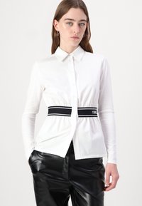 Camicia bianca a maniche lunghe con chiusura frontale a bottoni, cintura in vita cinchata nera con strisce bianche, indossata con pantaloni neri lucidi.