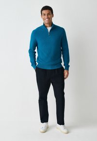 Türkiser strukturierter Pullover mit kurzer Reißverschluss-Kragen, kombiniert mit dunklen Hosen und weißen Sneakers. Glattes Material sowie gerippte Bündchen und Saum.