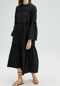 Robes noires à volants en tissu doux, avec des manches longues, des accents froncés et une coupe décontractée, associées à des sandales brunes à lanières.