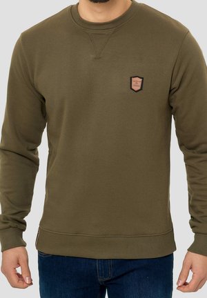 Homme portant un sweat-shirt col rond vert olive avec une petite écusson marron et noir sur la poitrine gauche, associé à un jean bleu foncé.
