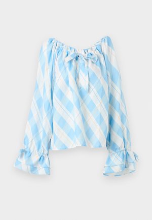Femme portant un chemisier à manches longues à carreaux bleu clair et blanc avec des liens aux poignets et au col, un jean bleu clair et des sandales beiges avec des nœuds.