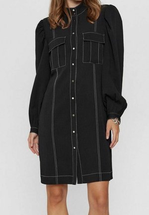 Robe noire avec boutonnage sur le devant, manches longues bouffantes et deux poches poitrine. Caractérisée par des coutures blanches visibles et une coupe décontractée.