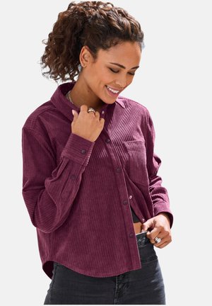 Frau mit lockigem Haar im Pferdeschwanz, die lächelt, eine violette Cord-Hemdjacke und dunkle Jeans trägt und dabei ihr Hemd richtet.