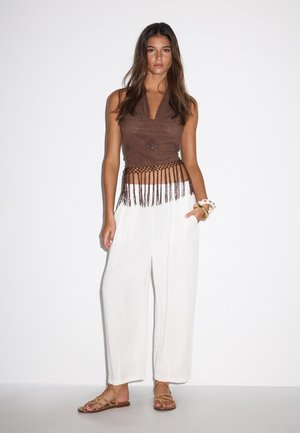 Jeune femme debout portant un haut marron sans manches avec des franges, un pantalon blanc à jambes larges, des bracelets dorés et des sandales beige sur fond blanc uni.