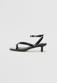 Sandale en cuir verni noir avec un talon bas, présentant des lanières croisées et une sangle de cheville ajustable. Surface lisse et brillante.